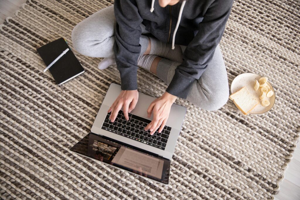 Woman in casual wear working remotely on a laptop, with a notebook and snack nearby.